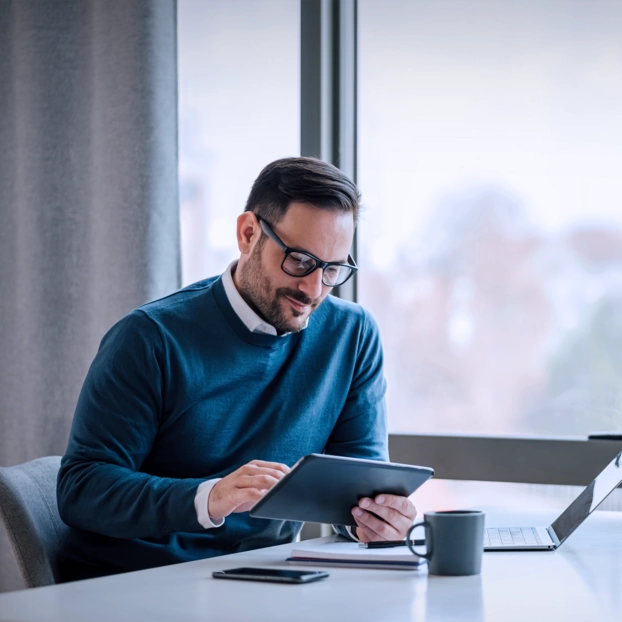 A man sits at a desk and uses the tablet for research. In the background you can see a large window with a view of a city.